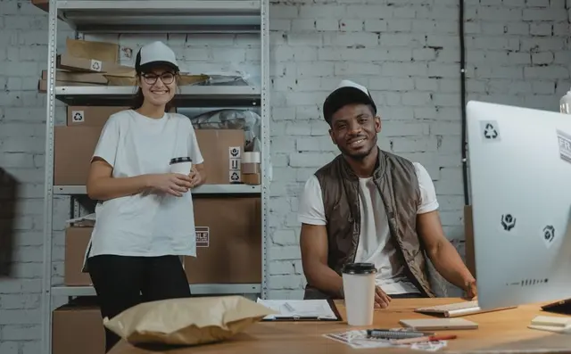 Two staff at a work table in a logistics space stacked with boxes